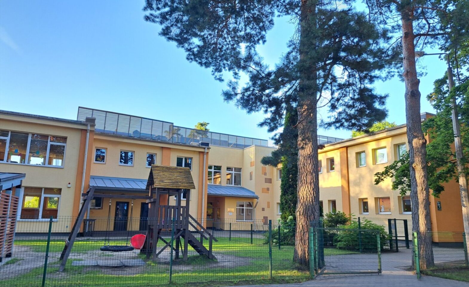 Dzeltena ēka, zemes un zāles seguma bērnu laukums, šūpoles, nojume.