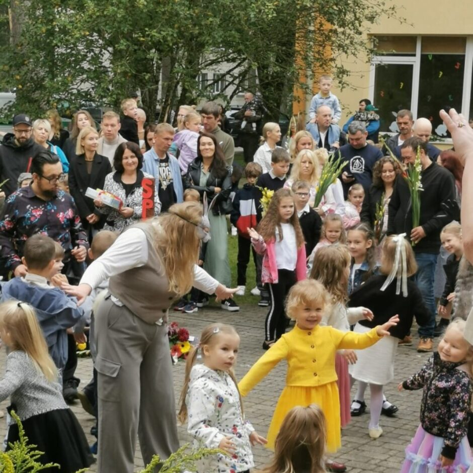 bērni un vecāki uz bruģa seguma pie dzeltenas ēkas.