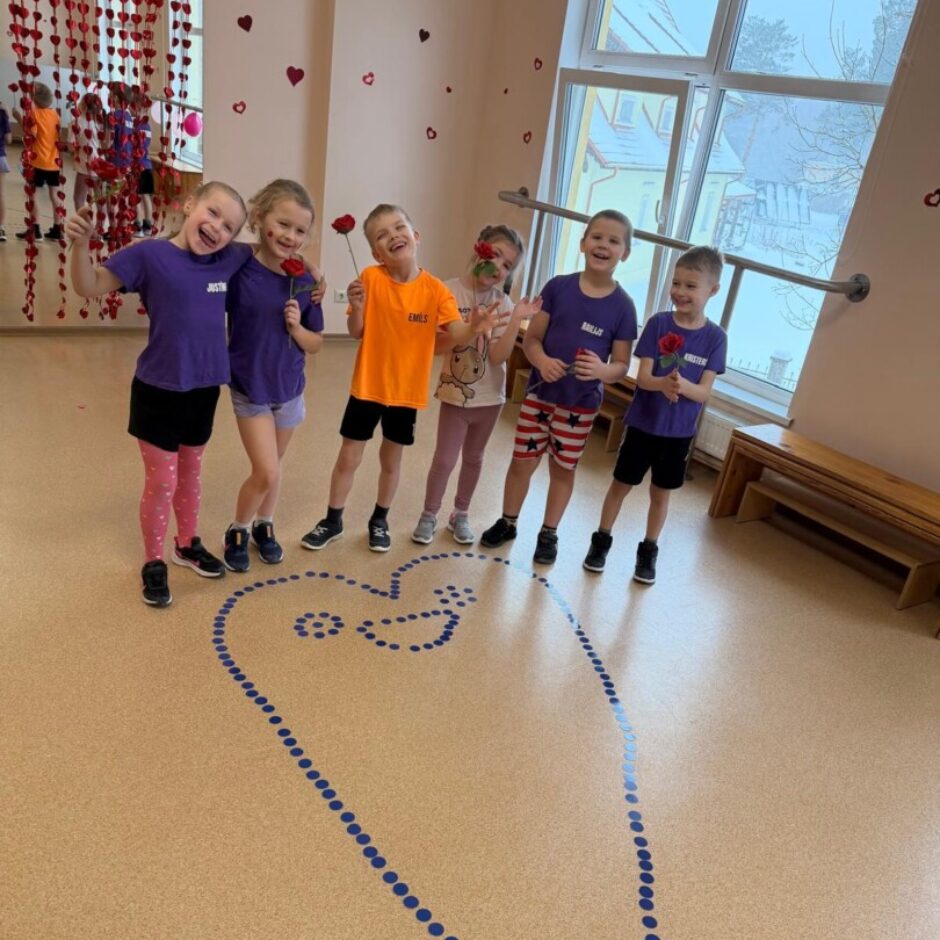 Uz brūnas grīdas izlikta zila sirsniņa. Aiz tās stāv bērni zilos krekliņos. Fonā zāles logs un sienas.