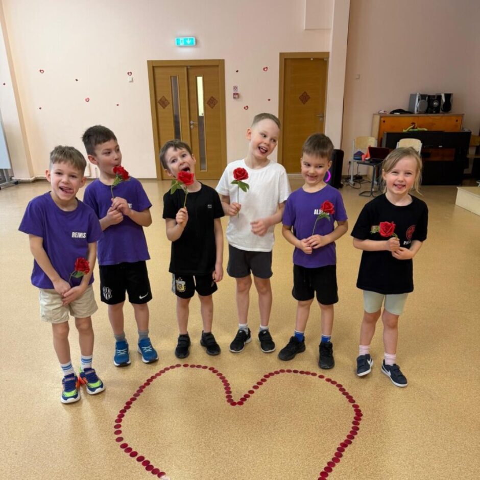 Uz brūnas grīdas izlikta sarkana sirsniņa. Aiz tās stāv bērni zilos krekliņos. Fonā zāles logs un sienas.
