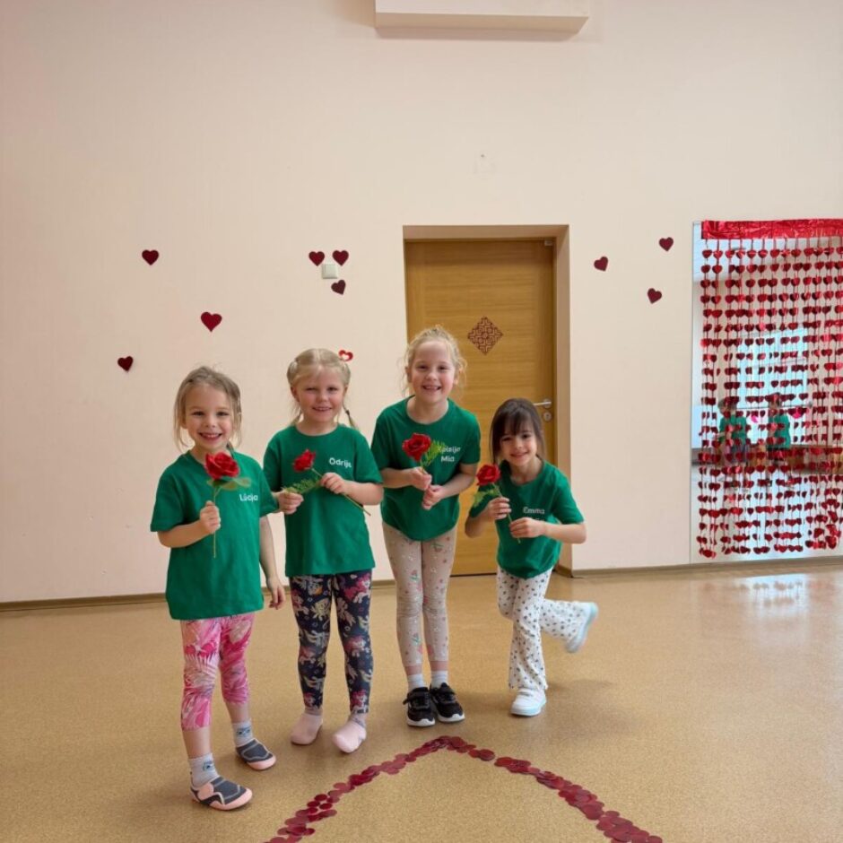 Uz brūnas grīdas izlikta sarkana sirsniņa. Aiz tās stāv bērni zaļos krekliņos. Fonā zāles logs un sienas.