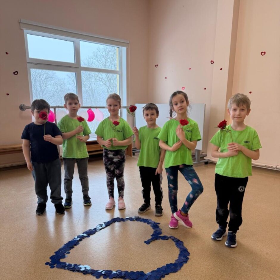 Uz brūnas grīdas izlikta zila sirsniņa. Aiz tās stāv bērni zaļos krekliņos. Fonā zāles logs un sienas.