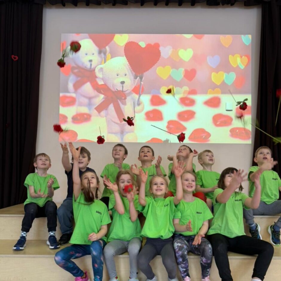 Priecīgi bērni zaļos krekliņos, rozes. Fonā ekrāns ar sirdīm un lāčiem.