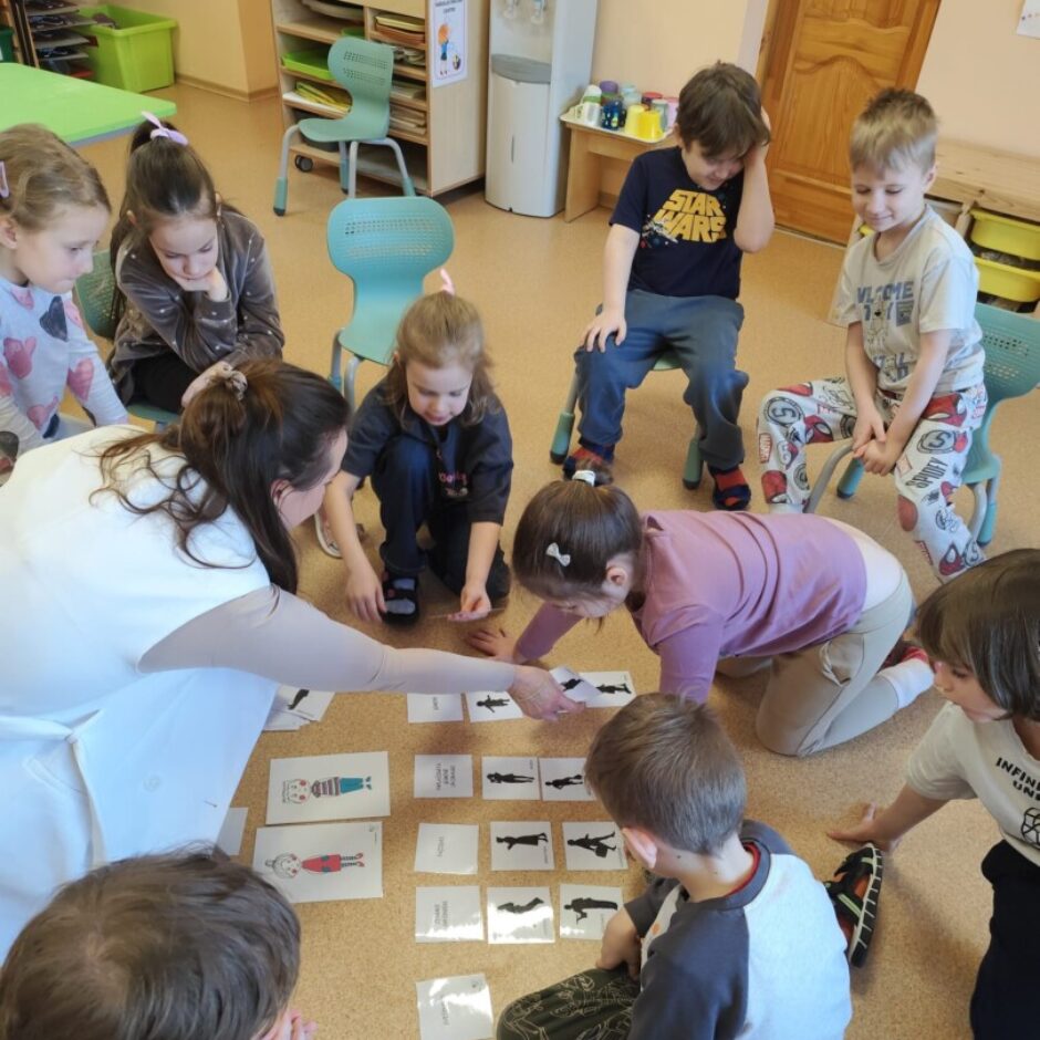 Skolotāja, bērni, Džimbas lelle. Grupas iekārtojums fonā.