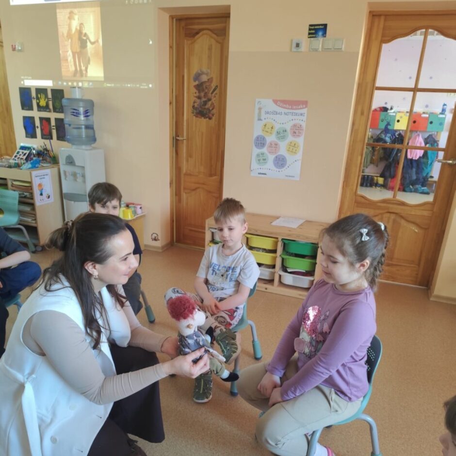 Skolotāja, bērni, Džimbas lelle, grupas iekārtojums fonā.