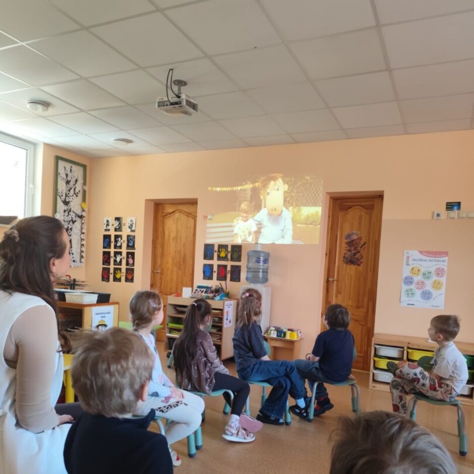 Gudrinieku grupas fons. Gaišas sienas, brūnas durvis. Bērni sēž uz krēsliem ar muguru. Bērni skatās ekrānā filmu.