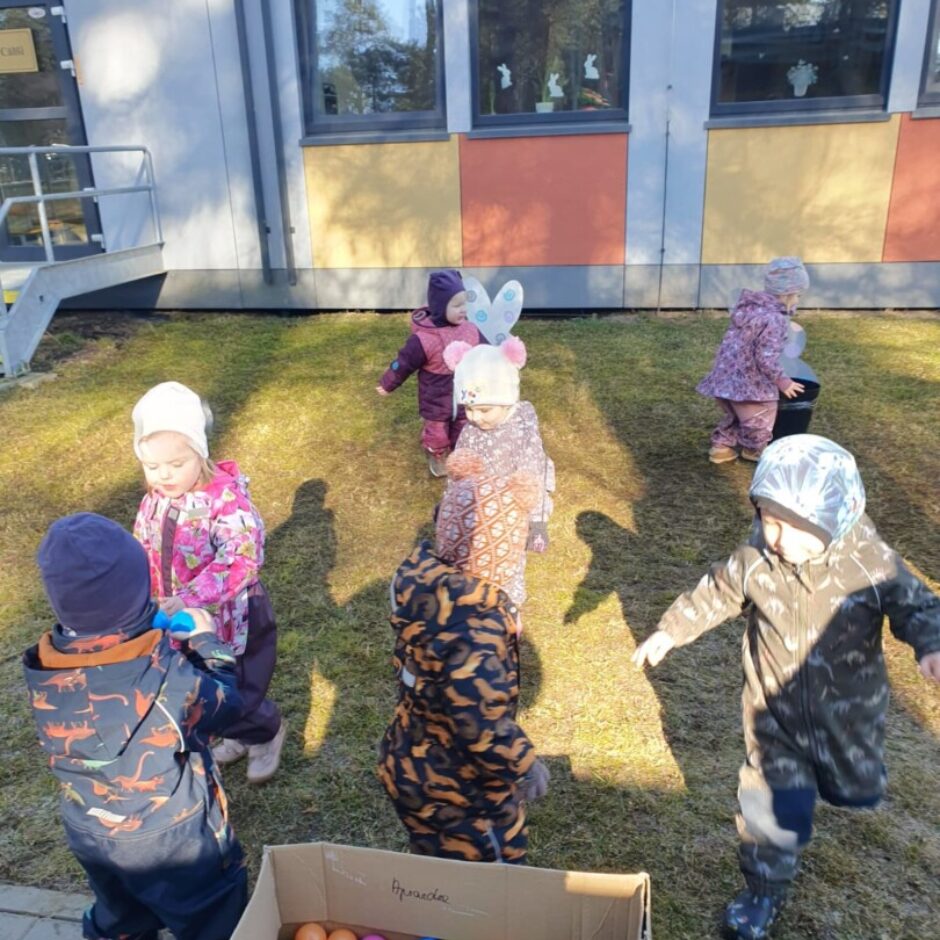 Vairāki mazi bērni spēlējas ārā saulainā dienā uz zāliena pie ēkas. Priekšplānā ir kartona kaste ar krāsainām bumbiņām.