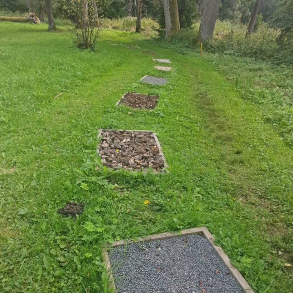 Baso pēdu taka ar virkni teksturētu posmu, kas pildīti ar granti, koka skaidām un tumšu augsni, vijas pa zaļu zālienu ar kokiem fonā.