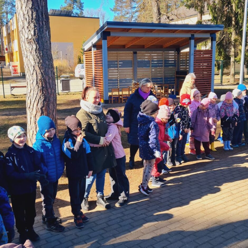 Liela bērnu grupa un vairāki pieaugušie stāv ārā saulainā dienā uz bruģēta laukuma. Bērni ir tērpušies cepurēs un jakās, viņi piedalās āra aktivitātē. Fonā redzami koki, koka nojume un ēkas.