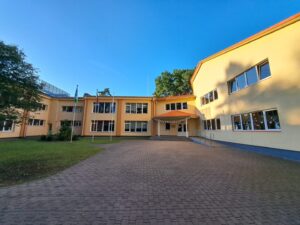 Gaiši dzeltens divstāvu skolas nams zem dzidrām zilām debesīm. Pie galvenās ieejas, ko sedz maza nojume un pie kuras ved uzbrauktuve, plīvo trīs karogi. Daži ēkas logi rotāti ar zaķu un tauriņu izgriezumiem.