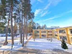 Dzeltena ēka sniegotā ainavā ar augstām priedēm, saulainā ziemas dienā. Pie ēkas plīvo Latvijas un citi karogi, bet tālāk redzama iela un tās malā bērni ar ragavām.