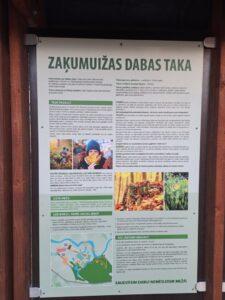 Informācijas stends ar uzrakstu "ZAKUMUIŽAS DABAS TAKA", kurā sniegta informācija par dabas taku, cilvēka maņām, sēņošanu un dabas saglabāšanu. Redzamas fotogrāfijas ar cilvēkiem dabā, sēnēm un augiem, kā arī takas karte. Stenda apakšdaļā ir sauklis "SAUDZĒSIM DABU! NEMĒSLOSIM MEŽĀ!".