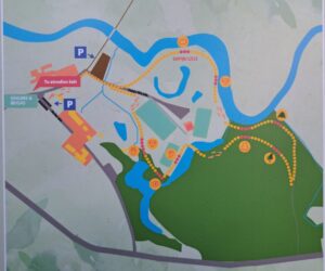 Dabas takas karte, kas attēlo maršrutu gar upi un caur parku ar ēkām. Uz kartes redzams uzraksts "Tu atrodies šeit" pie starta punkta, kas atzīmēts kā "SĀKUMS & BEIGAS" pie "Skolas iela" un "Sapņu lIča iela". Dzelteniem punktiem iezīmētā taka ved uz "Sapņu Līcis" un ietver sajūtu punktus. Ir norādītas arī autostāvvietas.