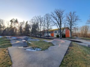 Tukša asfalta pumptrack trase ar dažādām rampām un virāžām uz zālāja, kurā redzami kūstoša sniega laukumi. Fonā liela balta un oranža ēka ar dzeltenīgu jumtu starp kailiem kokiem zem skaidrām zilām debesīm.