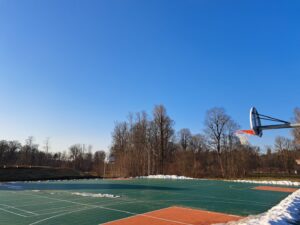 Āra basketbola laukums ar zaļu un oranžu segumu, kura malās redzami sniega pleķi. Fonā kaili koki zem dzidri zilas debess. Labajā pusē izcelts viens basketbola grozs ar oranžu tīklu, un otrs grozs redzams tālāk kreisajā pusē.