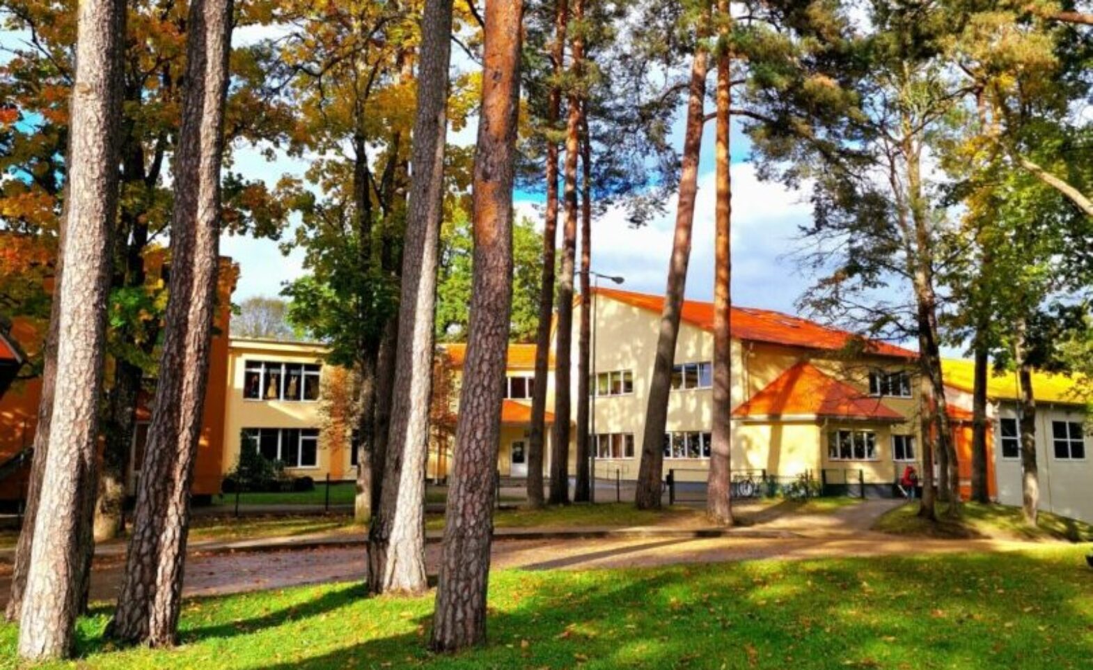 Rudens skats uz dzeltenu ēku ar oranžu jumtu, kuru ieskauj garas priedes un koki ar dzeltenām lapām, priekšplānā zaļš zāliens ar rozā ziediem.
