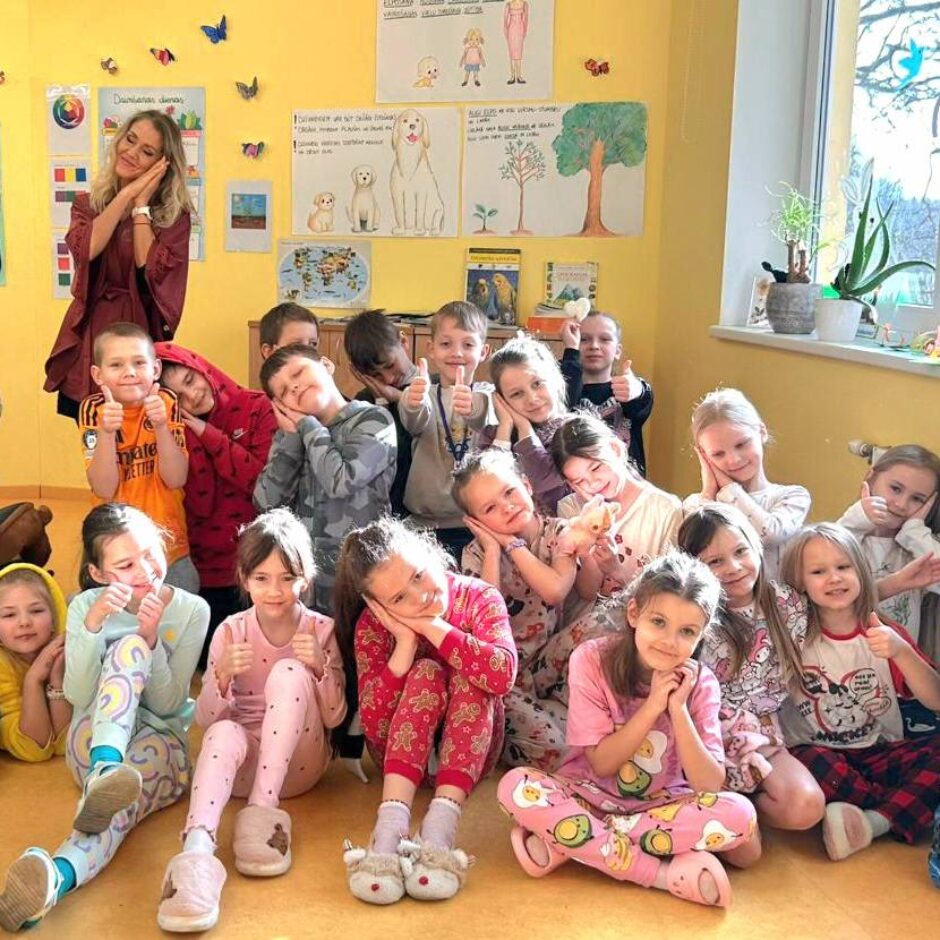 Skolotāja un aptuveni divdesmit bērni pidžamās pozē klases telpā. Lielākā daļa bērnu un stāvošā skolotāja tur rokas zem vaiga, atdarinot gulēšanu, kamēr daži bērni rāda īkšķus uz augšu.