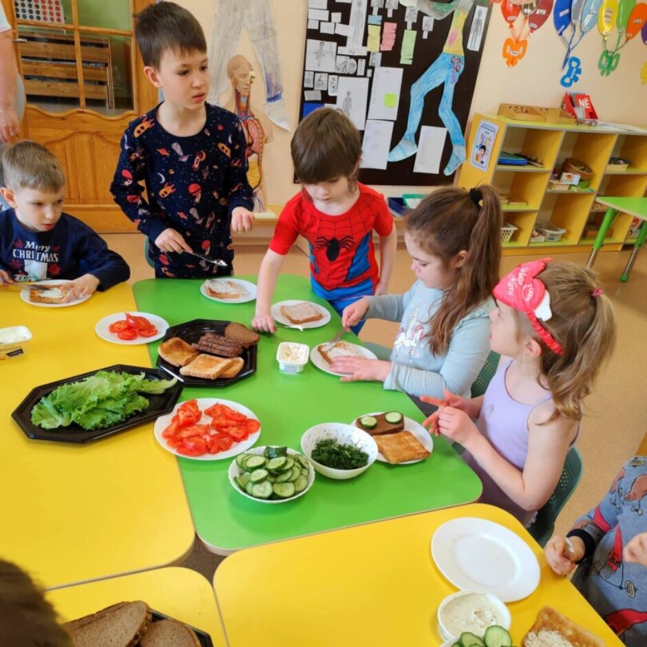 Bērni pie zaļiem un dzelteniem galdiem gatavo sviestmaizes ar maizi, smēriņiem un dārzeņiem, piemēram, salātiem, tomātiem un gurķiem.