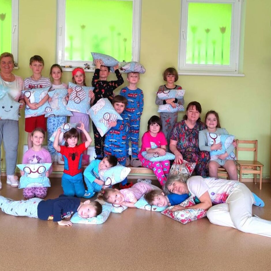 Bērnu un pieaugušo grupa ar spilveniem gaiši zaļā bērnudārza telpā. Daudzi bērni ir ģērbušies pidžamās, daži stāv, citi sēž vai guļ uz grīdas, turot dekoratīvus spilvenus.