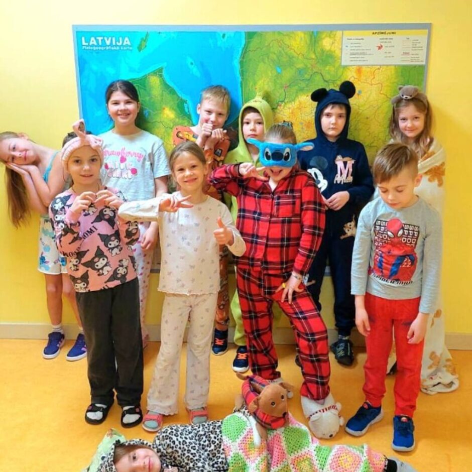 Desmit bērni pidžamās pozē fotogrāfijai. Aiz viņiem pie dzeltenas sienas ir liela karte ar uzrakstu 