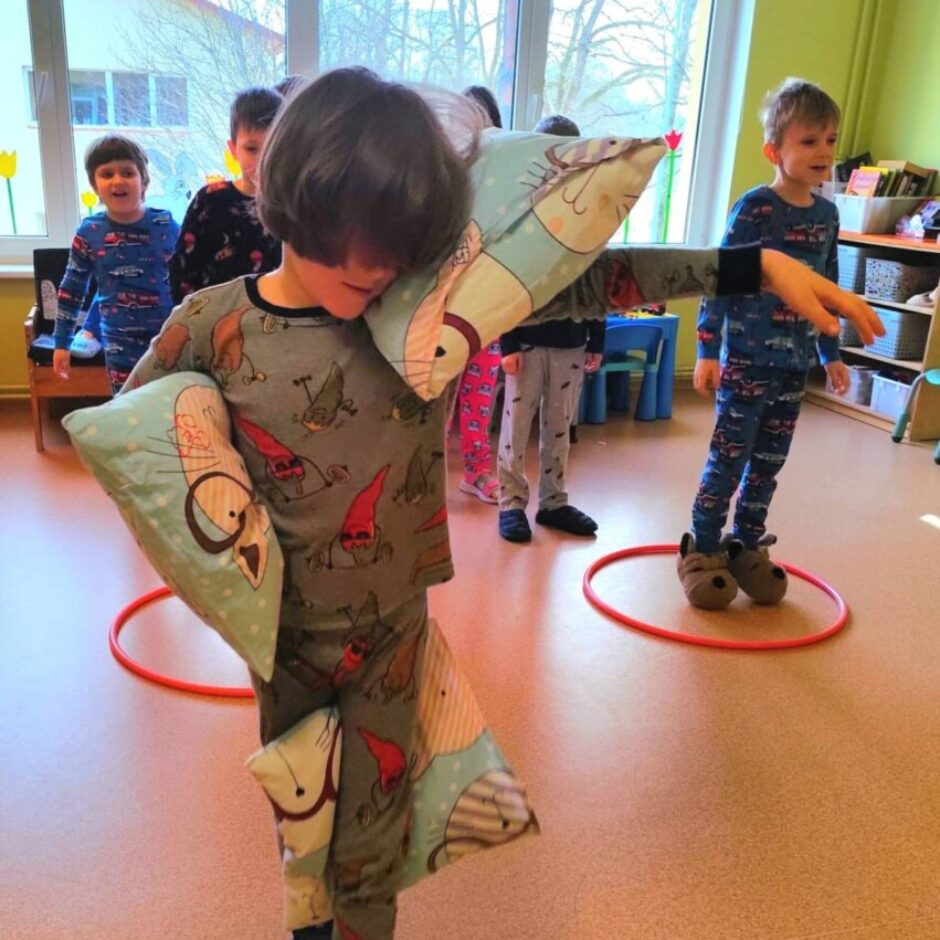 Bērns pelēkā pidžamā ar gnomu apdrukām tur un balansē ar trim maziem ziliem spilveniem, kas iespiesti zem rokām un pie kājas, ejot pa telpu. Fonā redzami citi bērni pidžamās, viens stāv sarkanā hula hopa aplī uz grīdas.