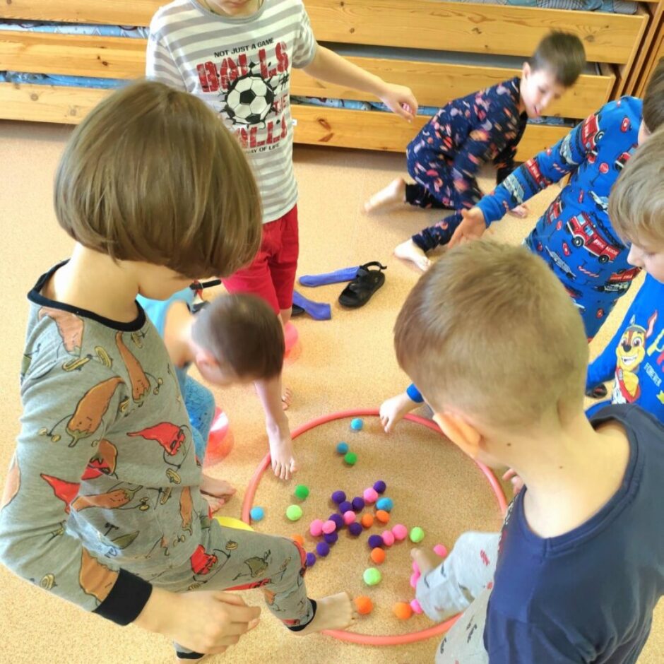 Vairāki bērni pidžamās rotaļājas uz gaiša paklāja telpā ar koka divstāvu gultām fonā. Viņi ir sapulcējušies ap rozā riņķi, kas piepildīts ar daudzām krāsainām pūkainām bumbiņām.