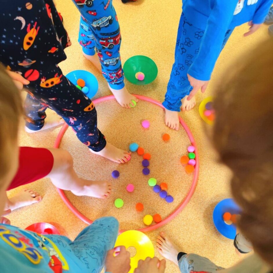 Vairāki bērni basām kājām stāv ap rozā hulahupu uz gaiši brūnas grīdas, ar pēdām mijiedarbojoties ar mazām, krāsainām bumbiņām, kas izkaisītas apļa iekšpusē. Blakus ir redzami citi rotaļu priekšmeti.