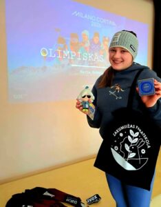 Smaidīga jauna sieviete, tērpusies sporta apģērbā, stāv pie projektora ekrāna, kurā redzami uzraksti "MILANO CORTINA 2026", "OLIMPISKĀS SPĒLES" un vārds "Marta Robeža" ar slēpotāju attēliem fonā. Viņa kreisajā rokā tur mazu lelli ar saulesbrillēm, bet labajā – zilu kastīti ar "SAMSUNG" un Olimpisko spēļu logotipiem. Uz viņas pleca ir melna auduma soma ar baltu logotipu un uzrakstu "ZAKUMUIŽAS PAMATSKOLA". Pie kājām redzams sporta inventārs ar uzrakstiem "Balcia insurance" un "uvex".