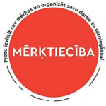 Sarkans aplis ar baltu tekstu “MĒRĶTIECĪBA”. Apkārt aplim ar melnu tekstu ir rakstīts: “Protu izvirzīt sev mērķus un organizēt savu darbu to sasniegšanai.”