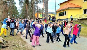 Liela bērnu grupa un daži pieaugušie ārā uz bruģēta ceļa veic sinhronus vingrinājumus ar paceltām rokām. Gar ceļu aug priedes, bet fonā redzama dzeltena skolas ēka.