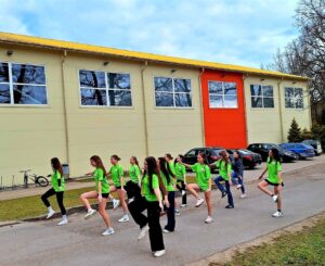 Vairākas meitenes spilgti zaļos T-kreklos vingro, ceļot ceļus, ārā uz asfalta ceļa. Fonā redzama gaiša sporta zāles ēka ar dzeltenu jumtu un oranžu sienas fragmentu, kā arī autostāvvieta.