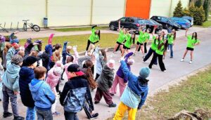 Āra pasākumā liela bērnu grupa aktīvi piedalās vingrinājumos, sekojot jauniešiem spilgti zaļos krekliņos, kas demonstrē kustības ar paceltām rokām un noliekšanos. Priekšplānā redzami jaunāki bērni neformālā apģērbā, bet fonā – gaiša ēka, autostāvvieta un koki.