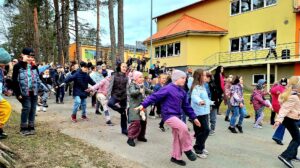 Liela bērnu grupa vingro ārā uz celiņa pie dzeltenas ēkas, daudzi līdzsvaro uz vienas kājas ar izstieptām rokām. Fonā redzami koki.