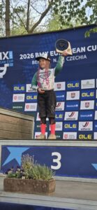 Jauna meitene ar rozā bizēm un cepuri stāv uz pjedestāla 3.vietas pakāpiena, smaidot un paceļot virs galvas trofeju. Viņa ir ģērbusies kreklā ar garām piedurknēm, šortos un rozā zeķēs. Aiz viņas ir zils reklāmas stends ar tekstu "2026 BMX EUROPEAN CUP Benátky nad Jizerou CZECHIA" un sponsoru logotipiem. Uz zemākā pjedestāla pakāpiena redzams skaitlis '3'.