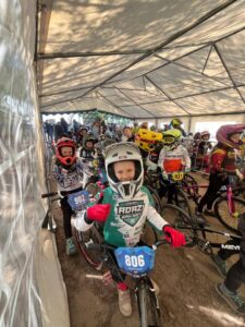 Priekšplānā smaidīga meitene baltā ķiverē un zaļi baltā kreklā ar uzrakstu "TEAM ADAZI BMX RACE" sēž uz BMX velosipēda ar zilu numura zīmi "806". Viņa valkā sarkanus cimdus. Aiz viņas teltī redzami vairāki citi bērni BMX ekipējumā ar velosipēdiem, tostarp viens ar numuru "902" un cits ar "931".