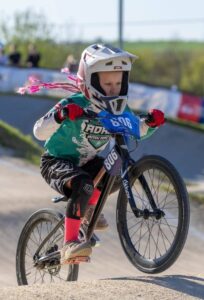 Jauna meitene baltā ķiverē ar rozā bizēm un sarkanos cimdos brauc ar BMX velosipēdu pa sacensību trasi. Uz velosipēda numura plāksnes un rāmja redzams numurs "806". Viņa valkā zaļu kreklu ar "ADAŽI BMX RACE" un melnus šortus ar ceļu sargiem.