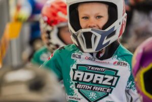 Jauna meitene baltā, zilā un sarkanā BMX ķiverē un tirkīzzilā sacīkšu kreklā, uz kura redzams logo "TEAM ADAZI BMX RACE", smaida un skatās tieši kamerā.