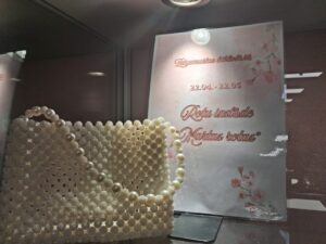 Balta pērļu rokassoma priekšplānā. Aiz tās redzama zīme ar tekstu: "Zaķumuižas bibliotēkā. 22.04. - 22.05. Rotu izstāde "Martas rotas"".