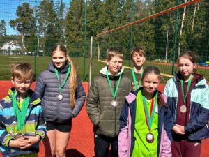 Seši bērni – trīs zēni un trīs meitenes – stāv uz sporta laukuma, katrs ar bronzas medaļu kaklā. Viņi smaida, fonā redzams sporta laukums un koki.