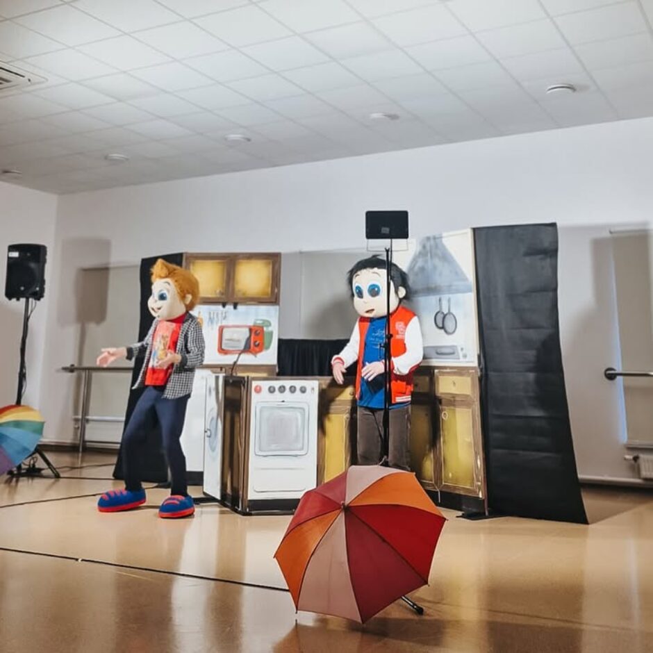 Divi aktieri multfilmu zēnu kostīmos uz skatuves, kas dekorēta kā virtuve. Viens tēls ar oranžiem matiem un rūtots krekls žestikulē pa kreisi, otrs ar tumšiem matiem un sarkanu vesti stāv labajā pusē. Uz virtuves dekorācijām redzama plīts, mikroviļņu krāsns ar uzrakstu 