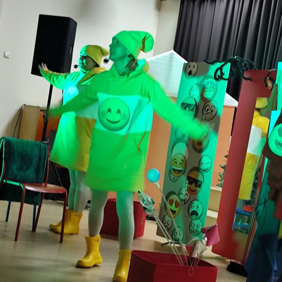 2 aktieri dzeltenās drēbēs stāv ar izstieptām rokām. Fonā skolas aktu zāle, spogules un stends ar dažādu emociju sejām.