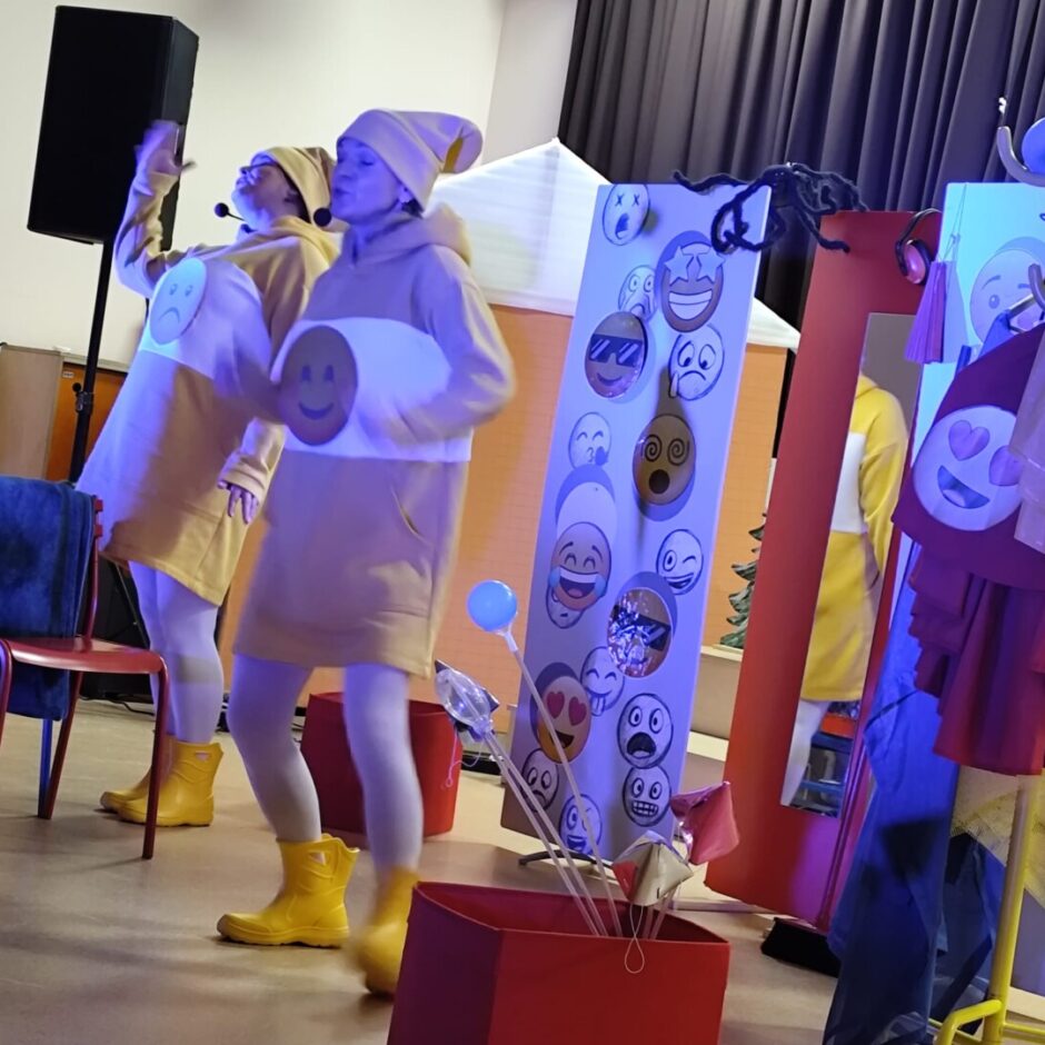 2 aktieri dzeltenās drēbēs, zābakos. uz skatuves fonā dekorācijas.