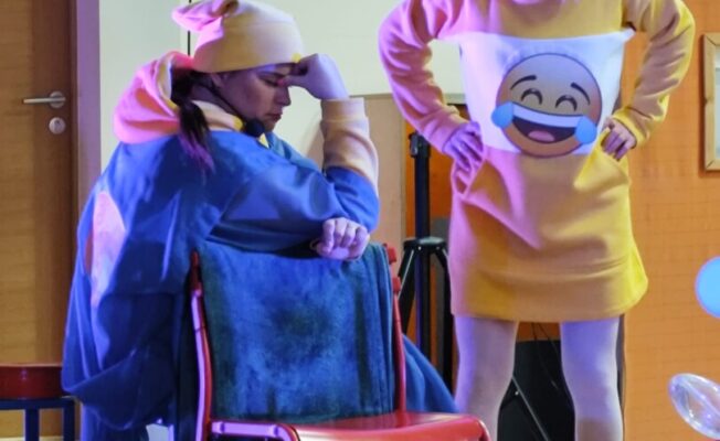 1 aktieris zilās drēbēs sēž, otrs - dzeltenās drēbēs stāv.