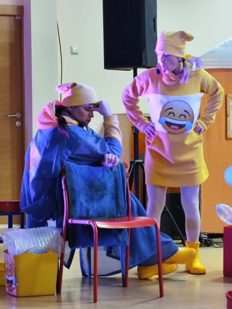 1 aktieris zilās drēbēs sēž, otrs - dzeltenās drēbēs stāv.