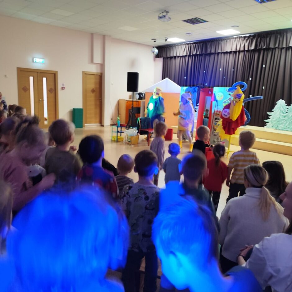 Bērni skatās izrādi. Fonā skatuve ar brūniem aizkariem un mākslinieki zaļās un dzeltenās drēbēs.