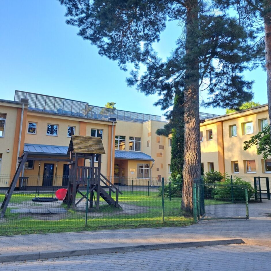 Dzeltena ēka, zemes un zāles seguma bērnu laukums, šūpoles, nojume.