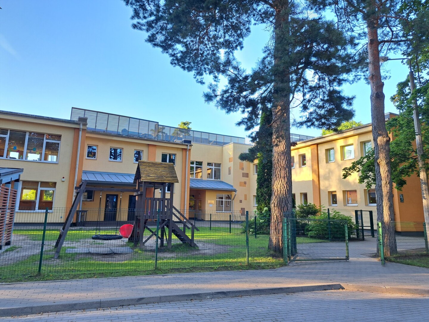 Dzeltena ēka, zemes un zāles seguma bērnu laukums, šūpoles, nojume.