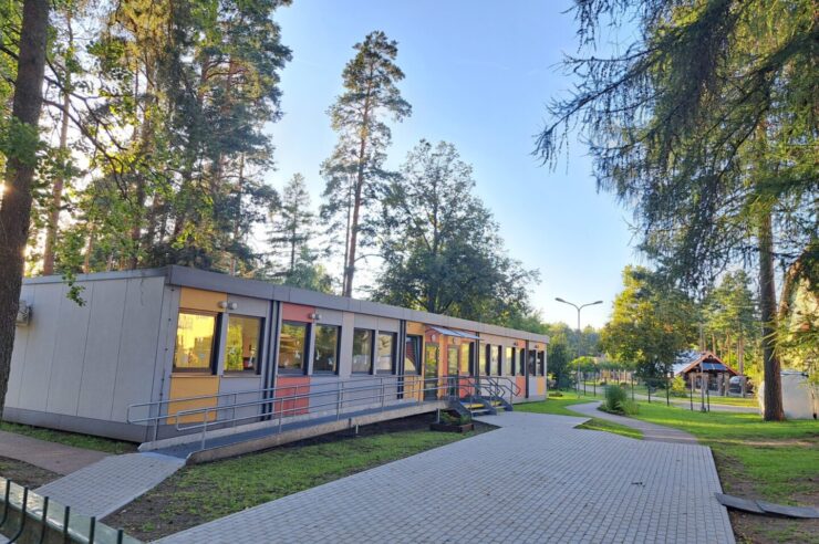 Pelēka moduļu ēka ar dzeltenu un oranžu dekorējumu. Priekšā bruģa segums un koki.