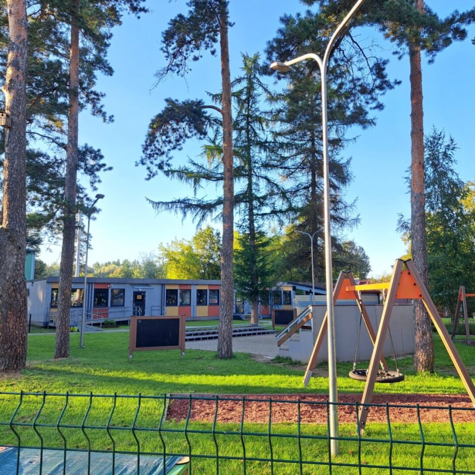 Pelēka moduļu ēka ar dzeltenu un oranžu dekorējumu. Priekšā bruģa segums un koki. Estrāde, bērnu laukuma šūpoles, zāles segums un mulčas segums.