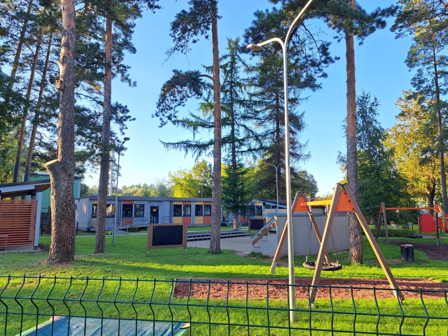 Pelēka moduļu ēka ar dzeltenu un oranžu dekorējumu. Priekšā bruģa segums un koki. Estrāde, bērnu laukuma šūpoles, zāles segums un mulčas segums.